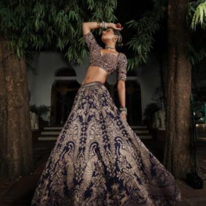 Woman wearing a deep purple raw silk lehenga with heavy gold zardozi embroidery featuring peacock motifs and matching elbow-sleeve blouse, posing outdoors at night