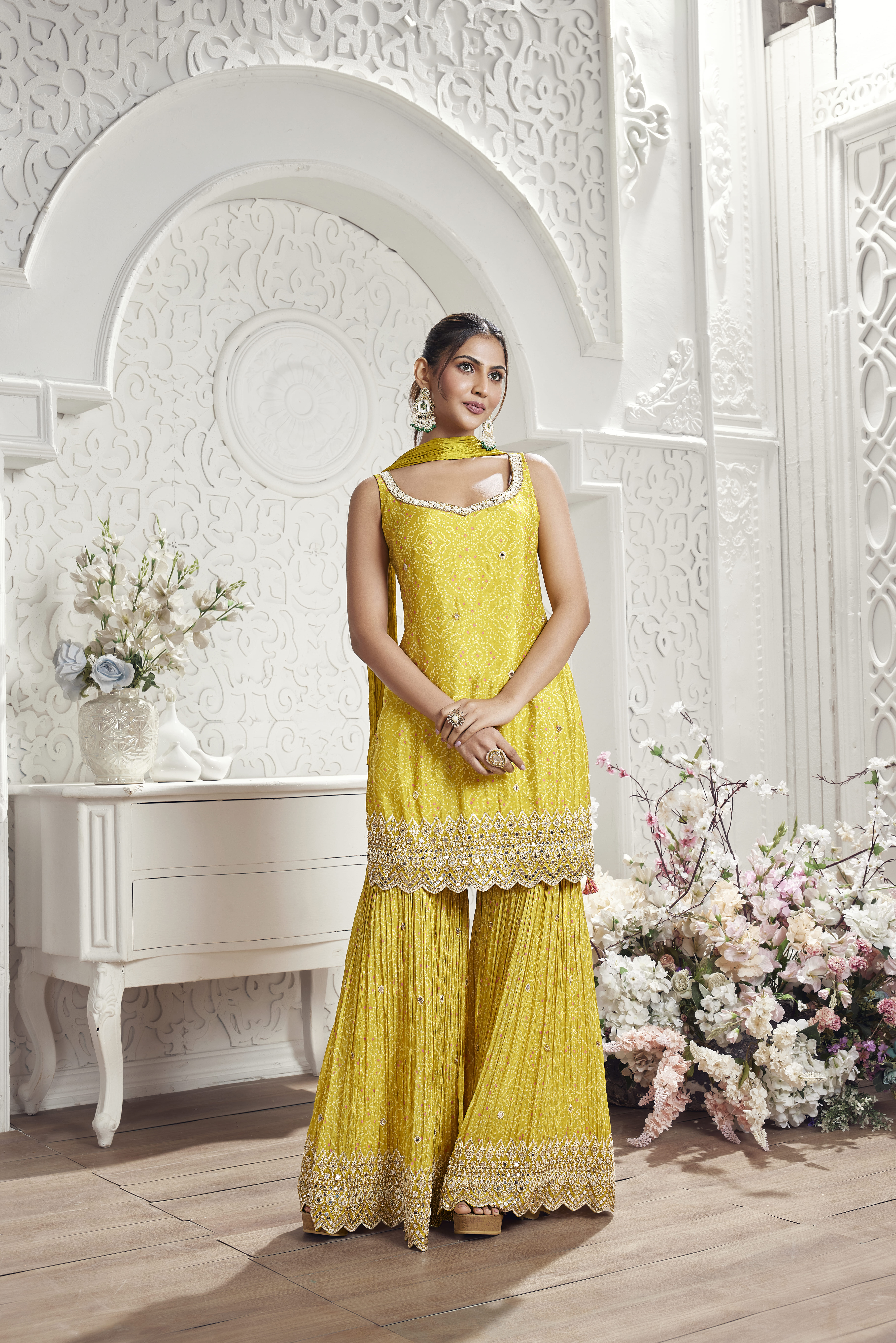 Model wearing a yellow embroidered sharara set with a sleeveless kurta and matching dupatta in an elegant floral-inspired interior setting.