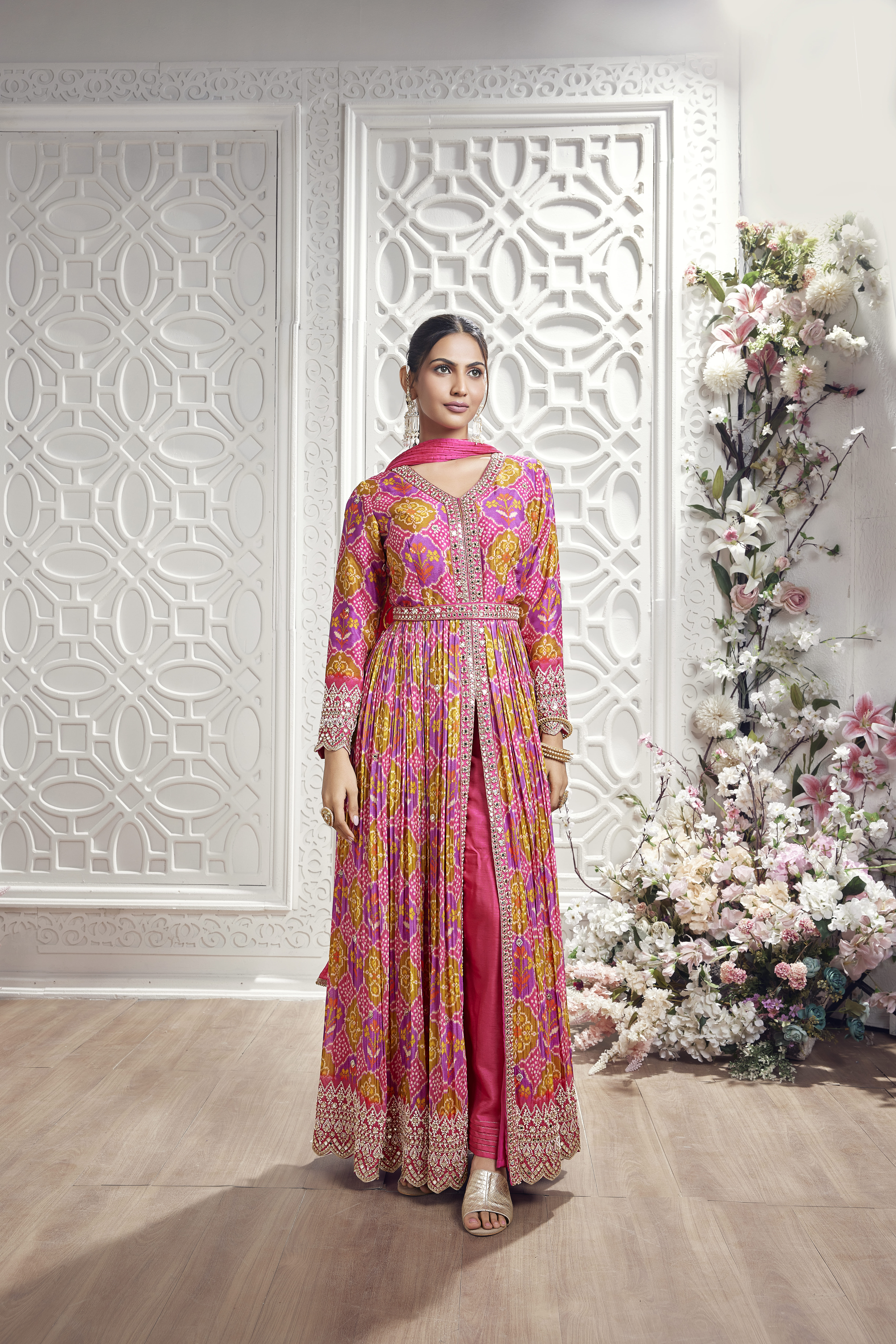A woman modeling a vibrant rani pink and mustard yellow Bandhani print jacket-style Anarkali suit with a front slit, embellished waist belt, and heavily embroidered scalloped borders, standing in front of a white carved decorative screen.