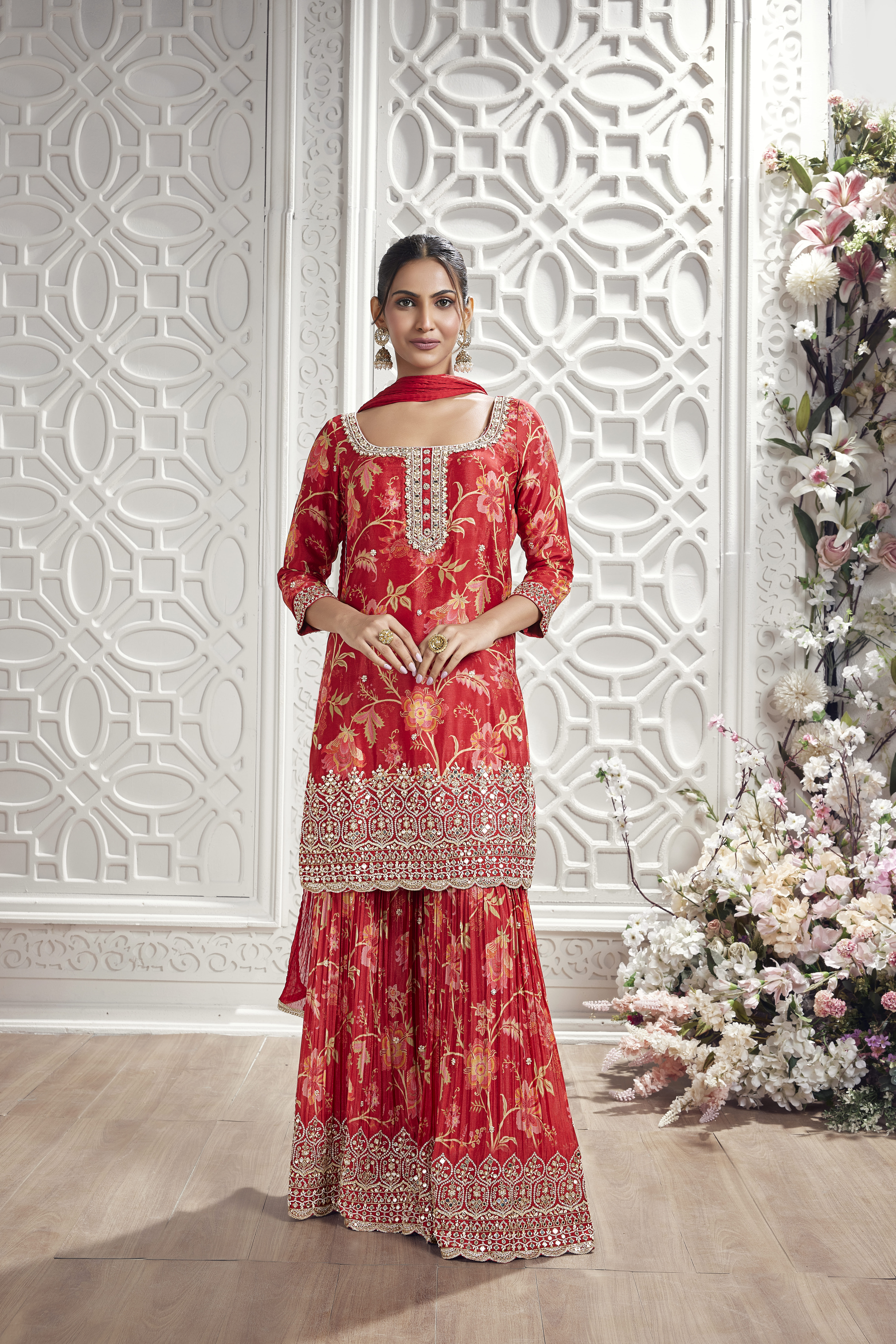Model wearing a crimson red floral embroidered sharara suit with matching dupatta in an elegant white floral interior setting.