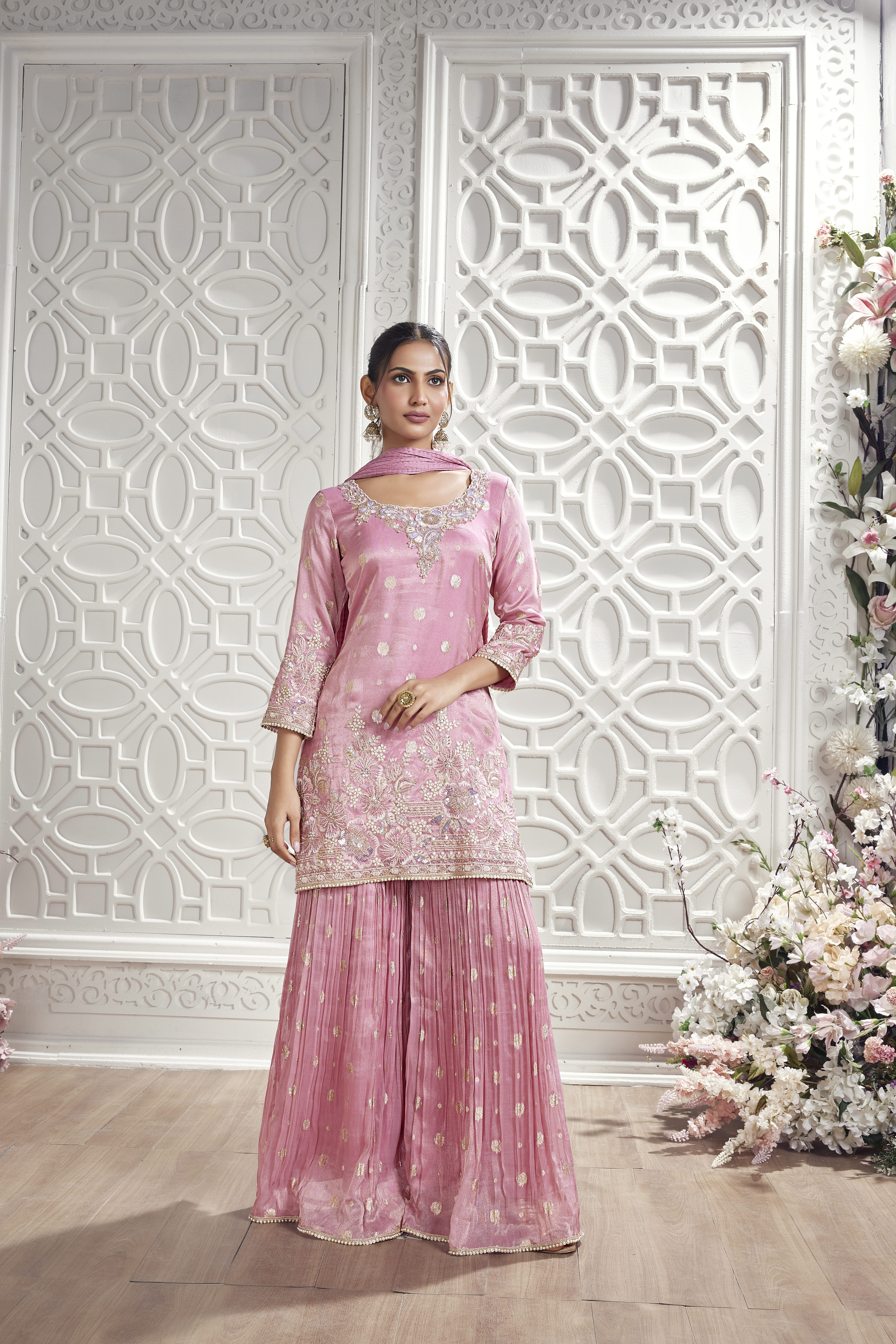 Model wearing a blush pink embroidered sharara suit with floral embellishments and matching dupatta in an elegant white interior setting.
