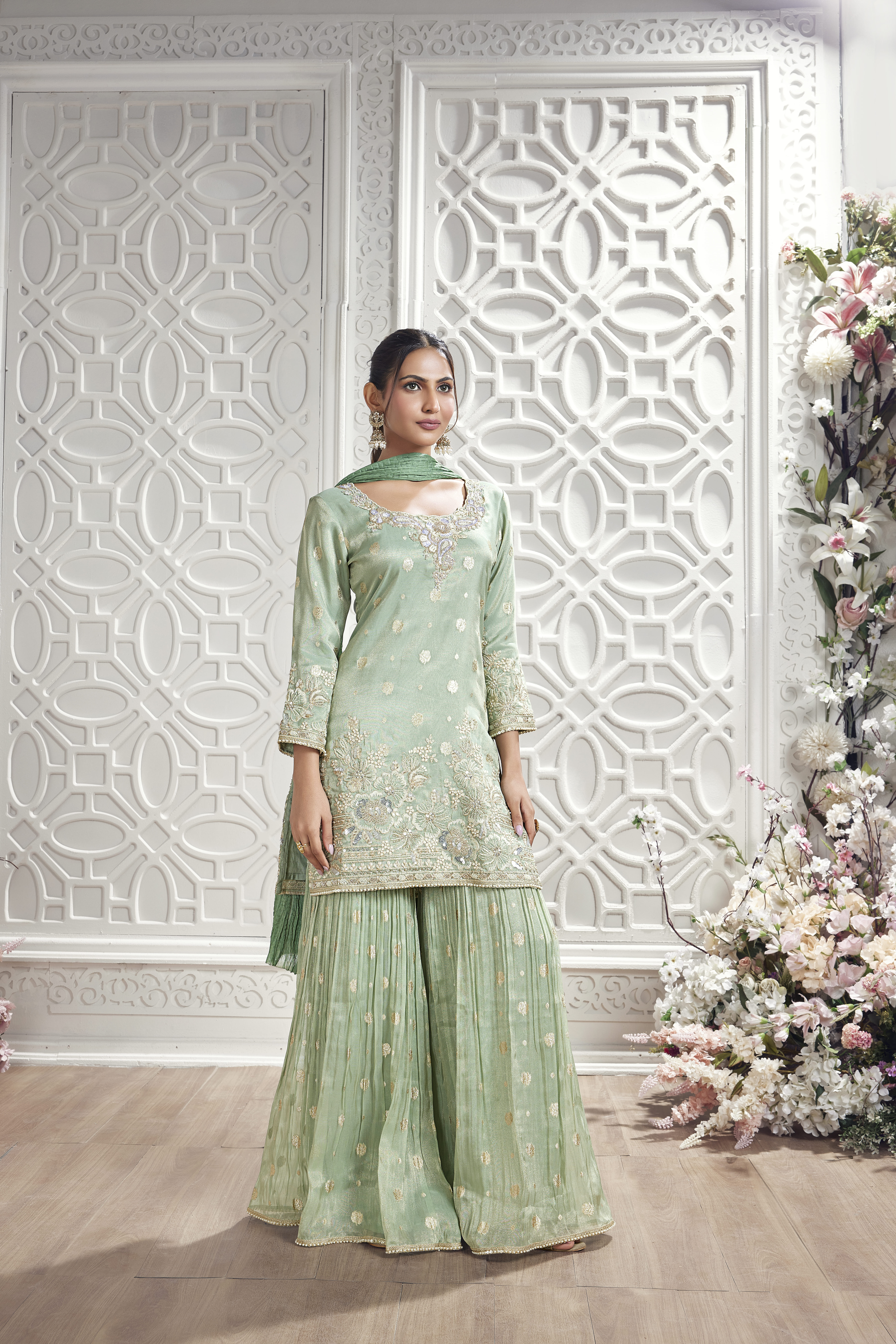 Woman wearing a mint green embroidered sharara suit with matching dupatta and pleated sharara pants in a luxurious white floral interior setting.