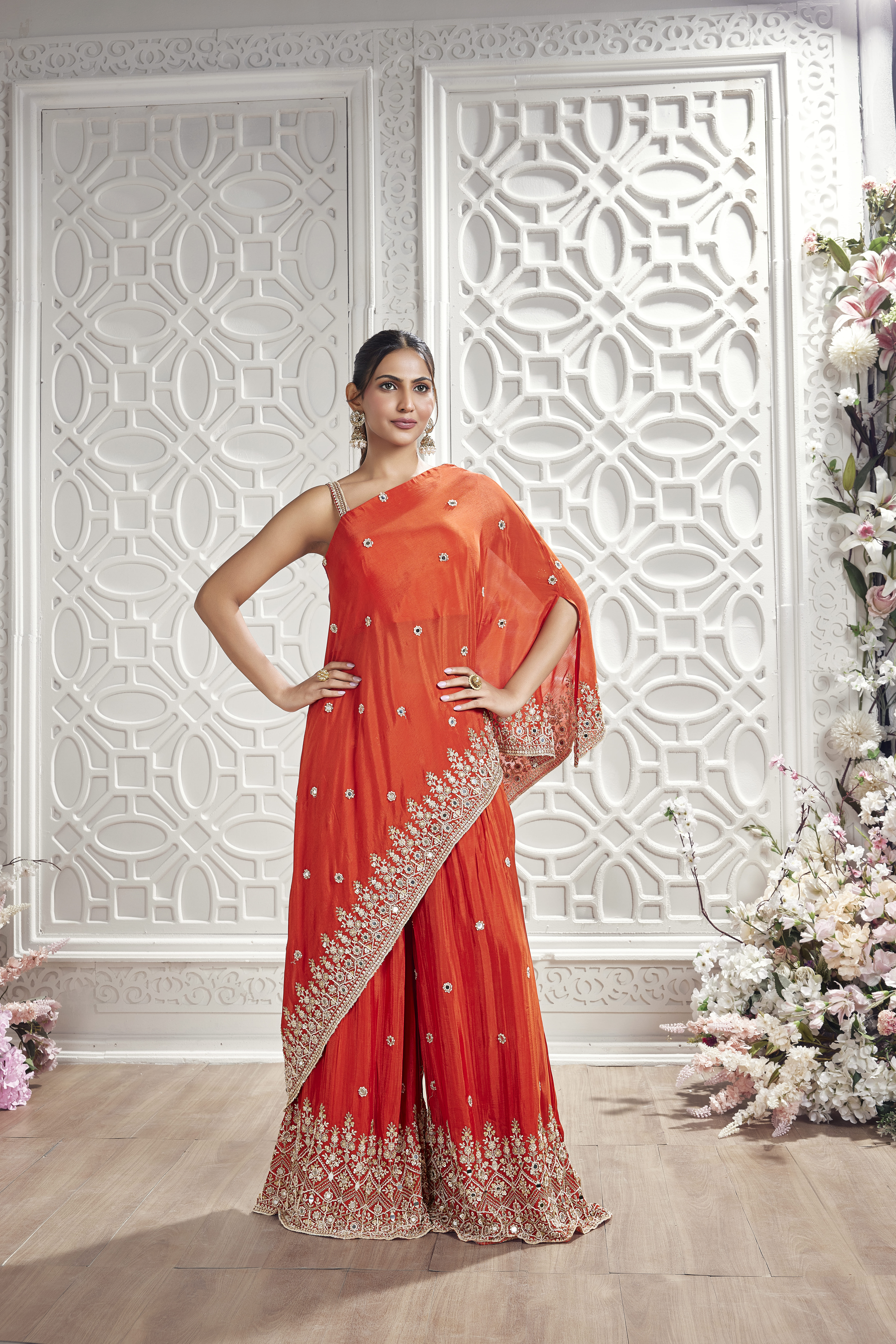 Woman wearing an orange embroidered pre-draped sharara saree with ornate border detailing and embellished sharara pants in an elegant white interior setting.