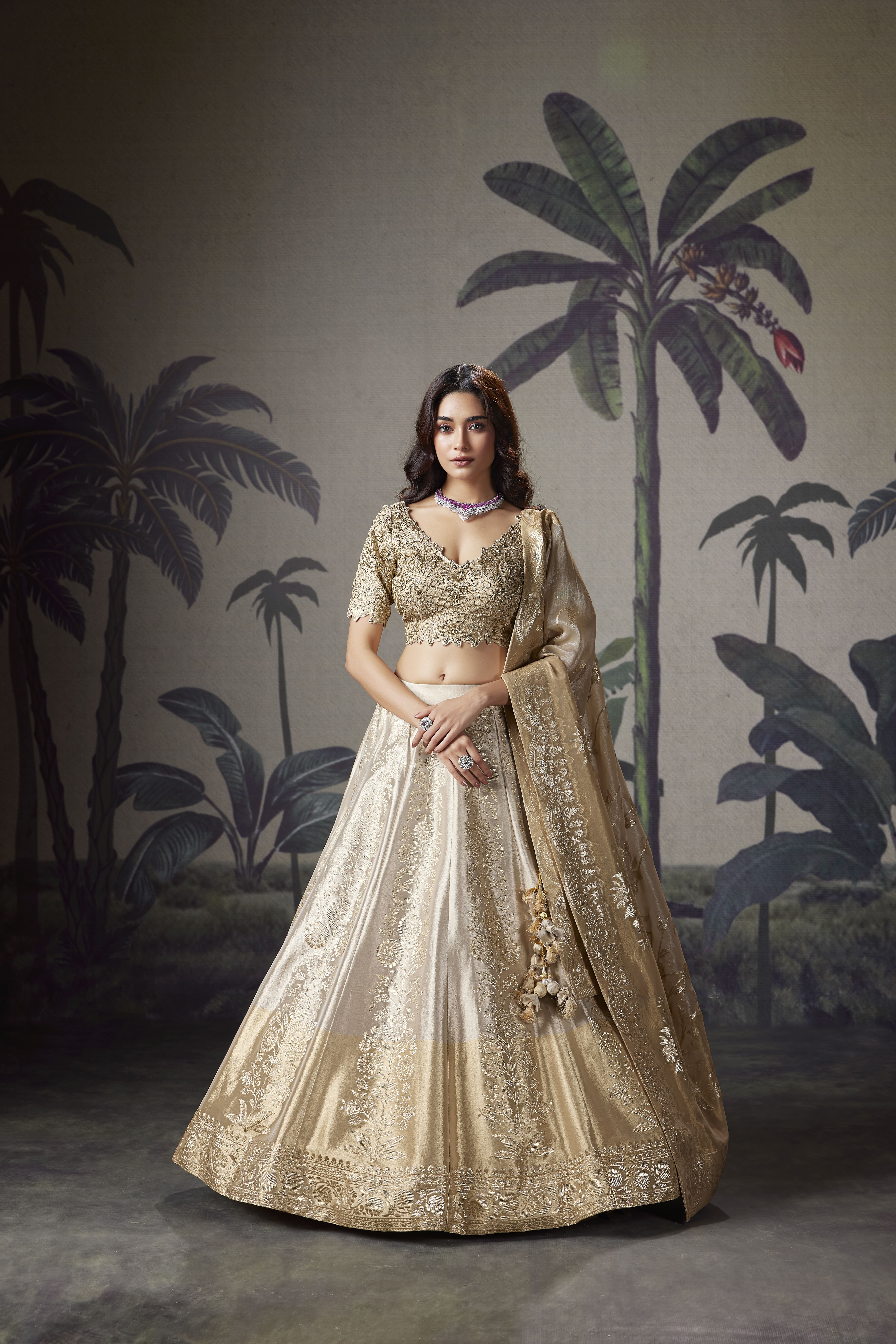 Woman wearing an ivory embroidered lehenga with a heavily embellished blouse and matching embroidered dupatta against a tropical-inspired backdrop.