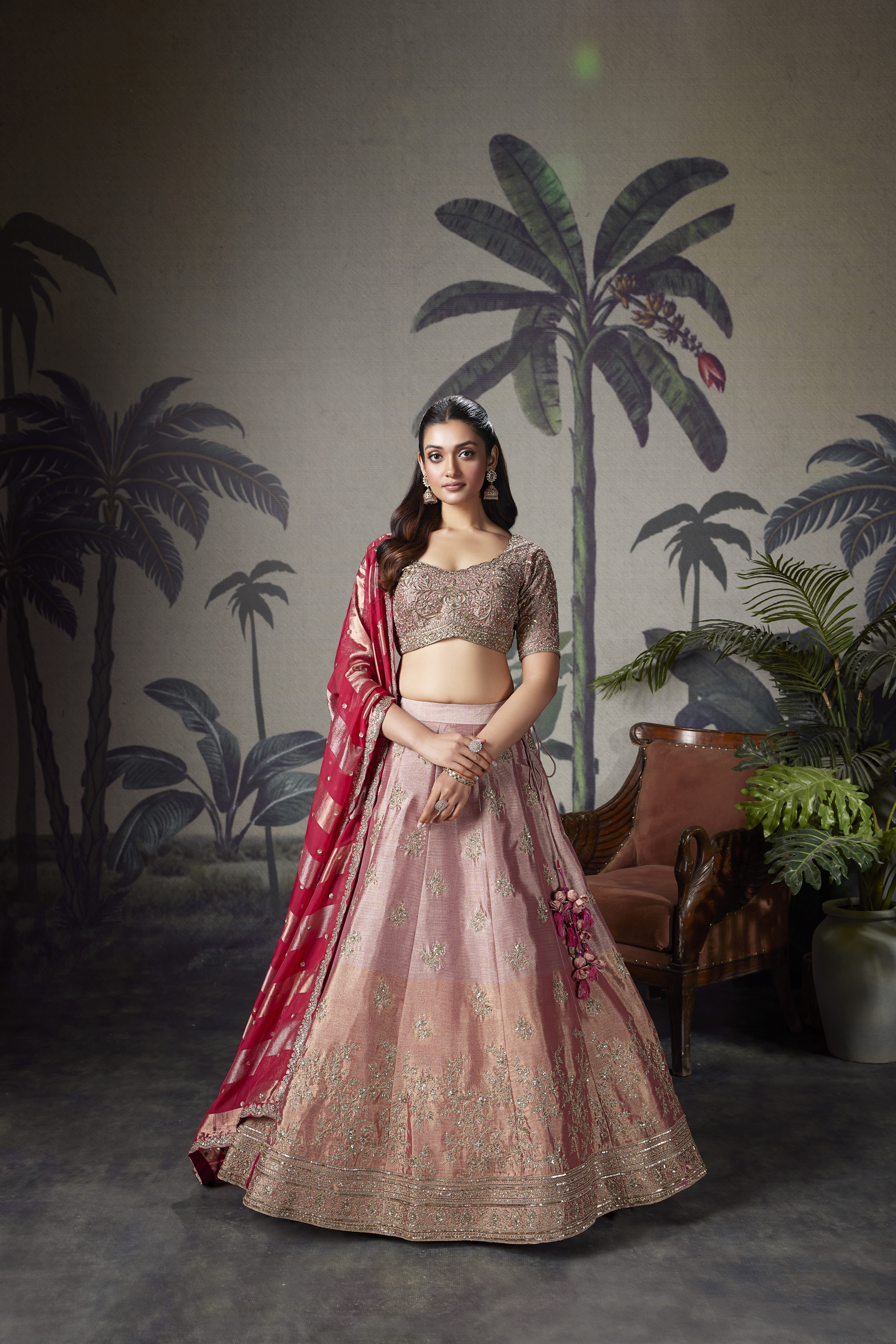 Model wearing a blush pink embroidered lehenga with a heavily embellished blouse and contrasting ruby pink dupatta, styled in an elegant indoor setting.