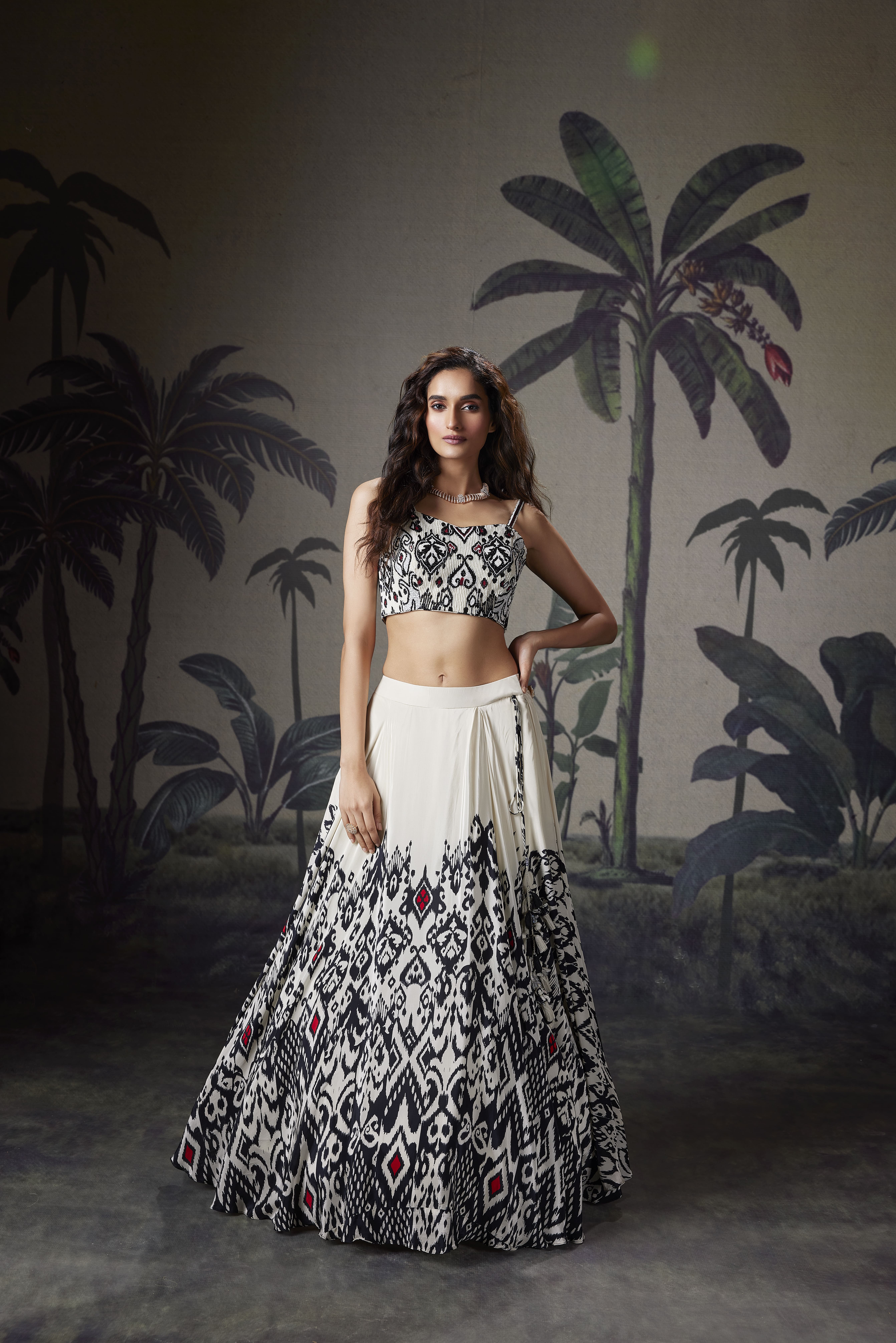 Model wearing an ivory lehenga set with black ikat-inspired prints and a matching sleeveless blouse, styled in a tropical-themed indoor setting.