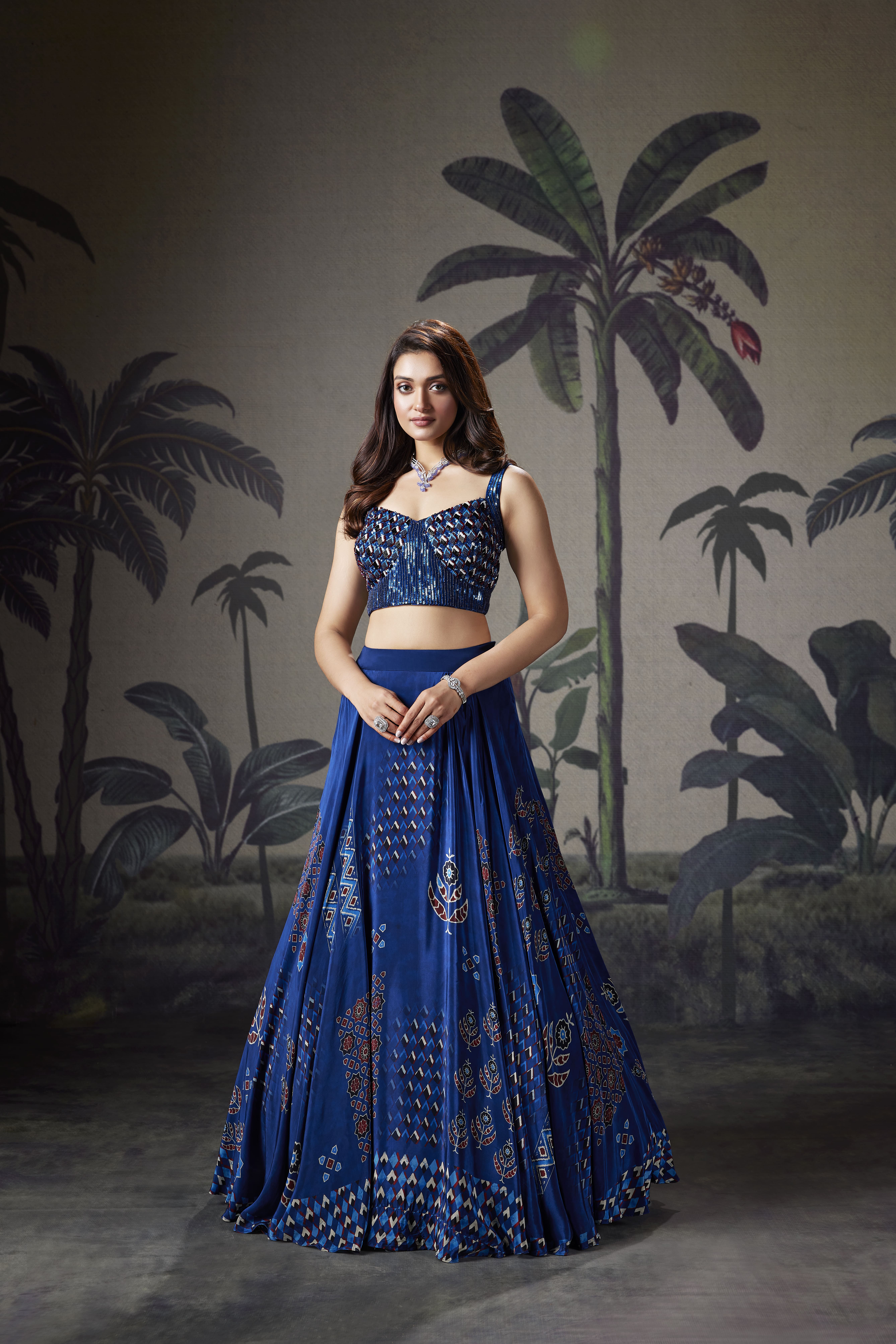 Model wearing a royal blue printed lehenga with an embellished sleeveless blouse, styled in an elegant tropical-inspired indoor setting.