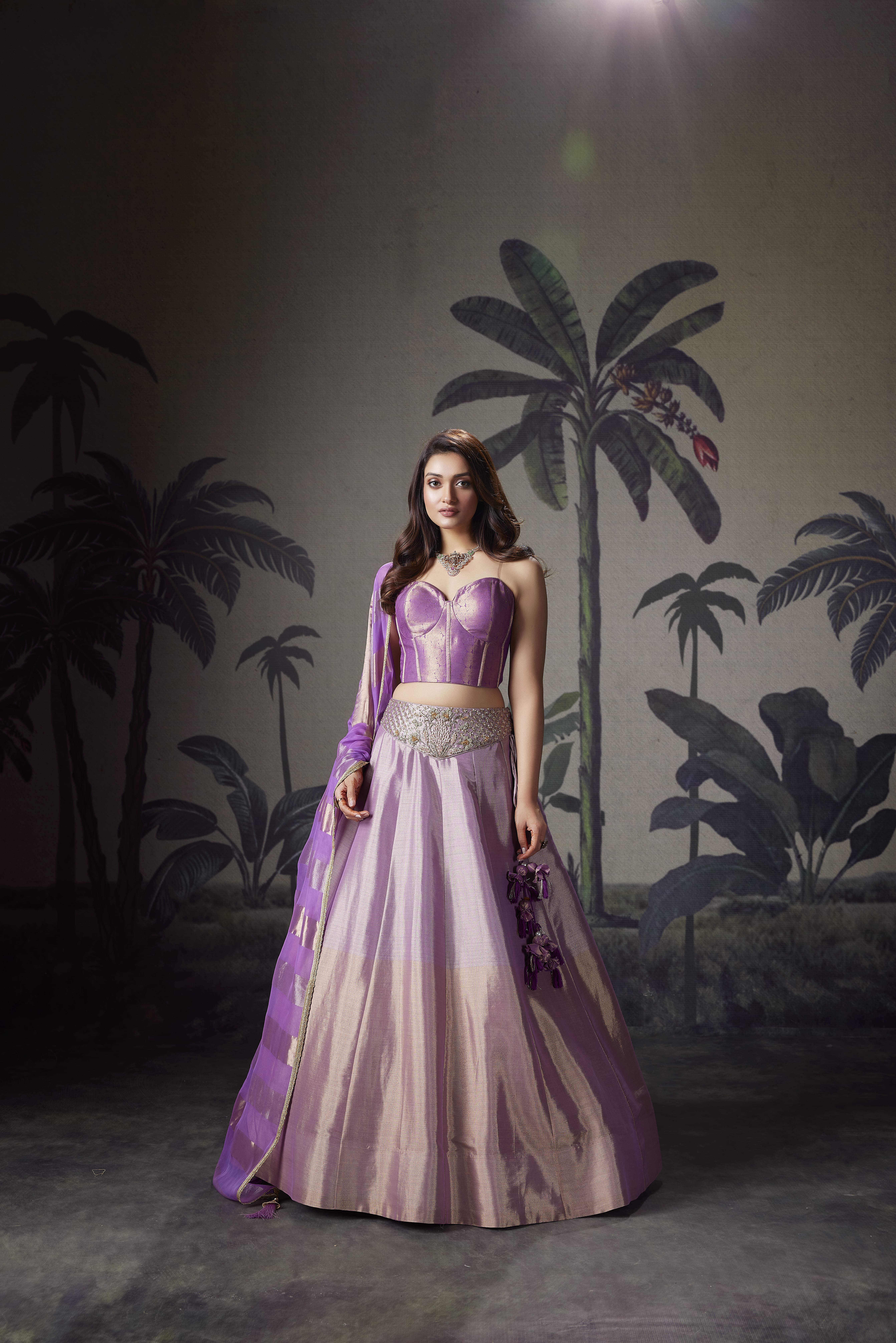 Model wearing a lilac corset blouse with a matching silk lehenga and sheer dupatta, styled in a tropical-inspired indoor setting.