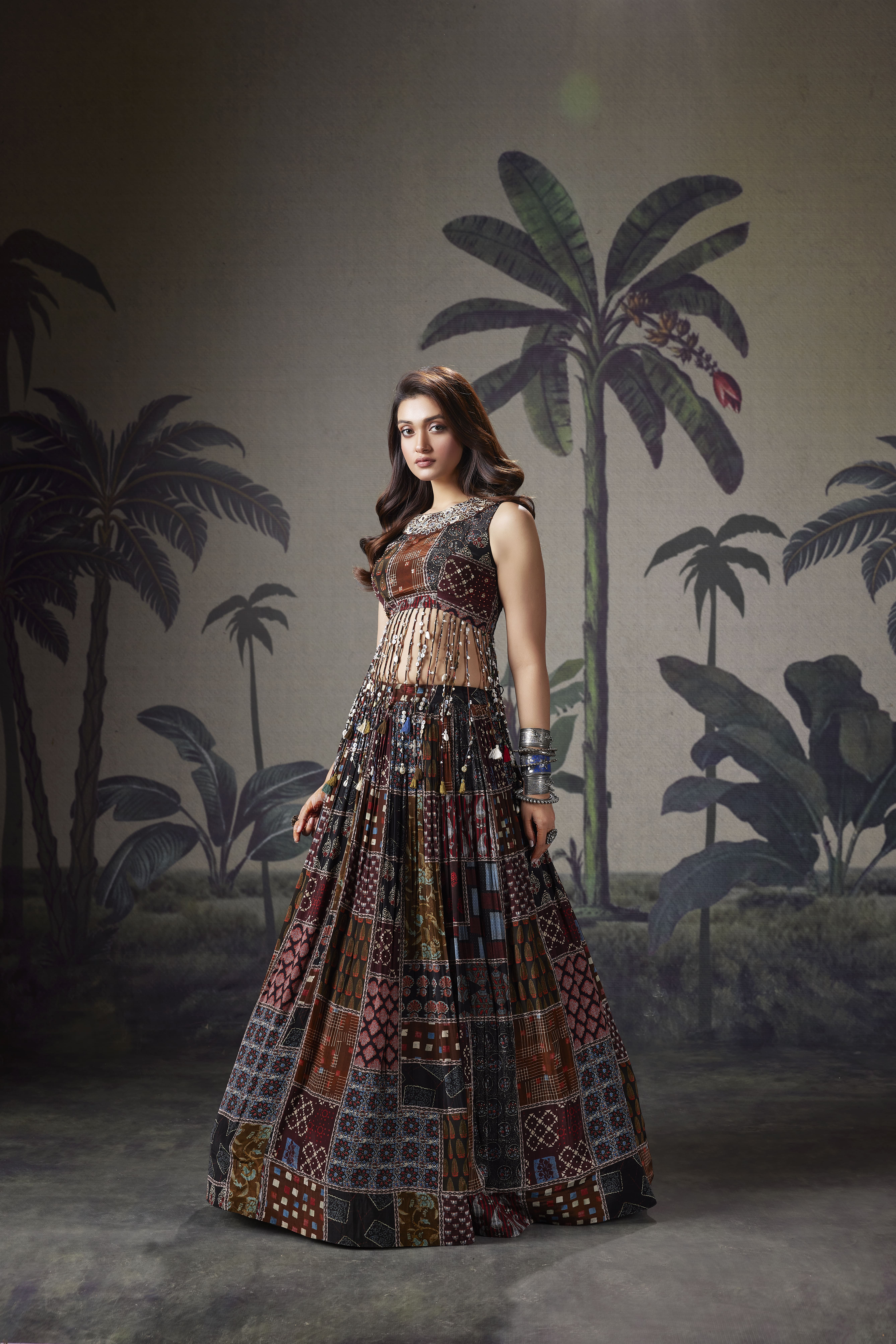 A woman posing in a multi-colored patchwork print lehenga skirt and a matching sleeveless blouse featuring a long fringe of mirror-work tassels and an embroidered neckline, set against a tropical palm tree backdrop.