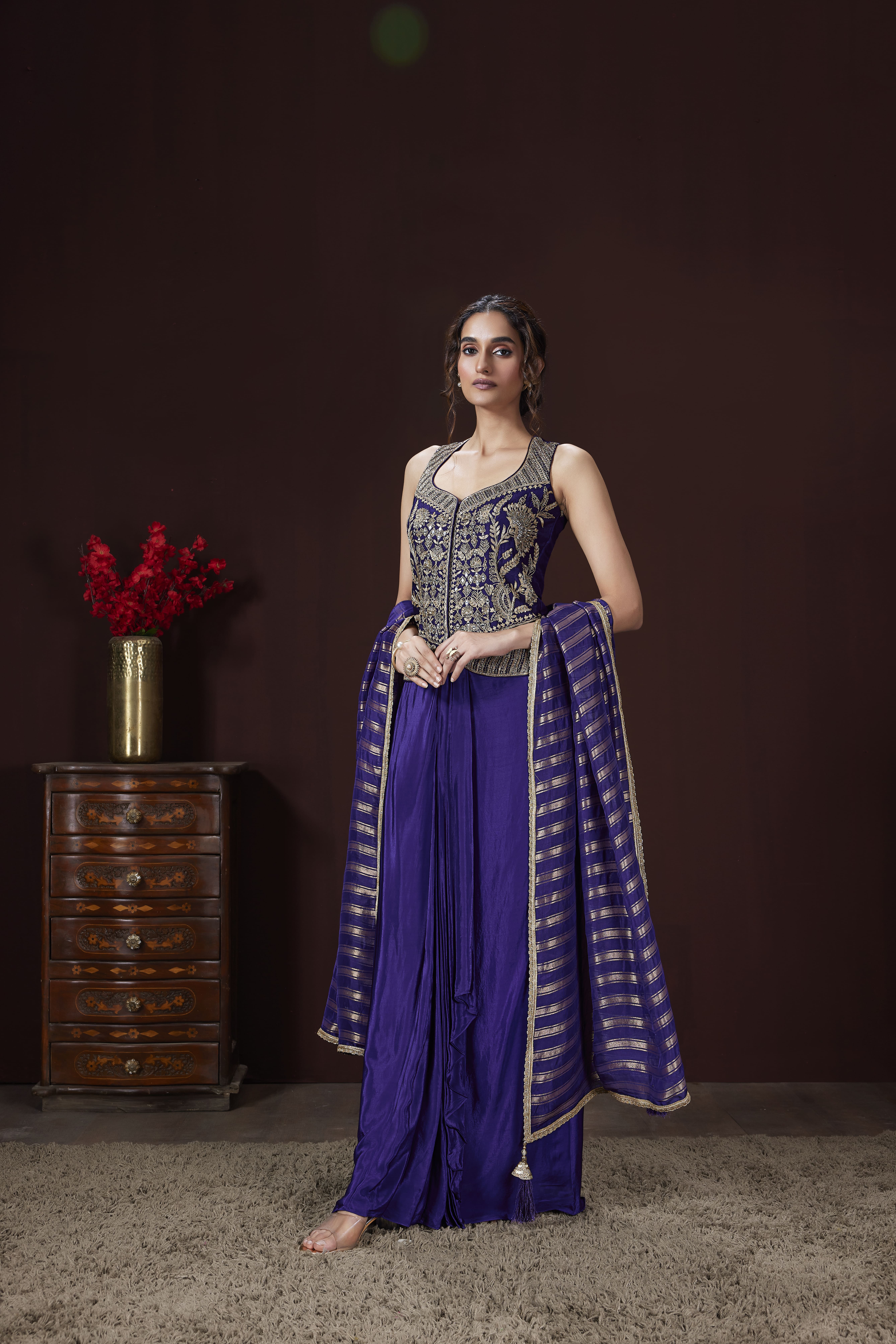 A woman modeling a regal violet Indo-Western fusion dress featuring a heavily gold-embroidered sleeveless corset top, a draped dhoti-style skirt, and a gold-striped dupatta, standing in a studio next to a vintage wooden cabinet.