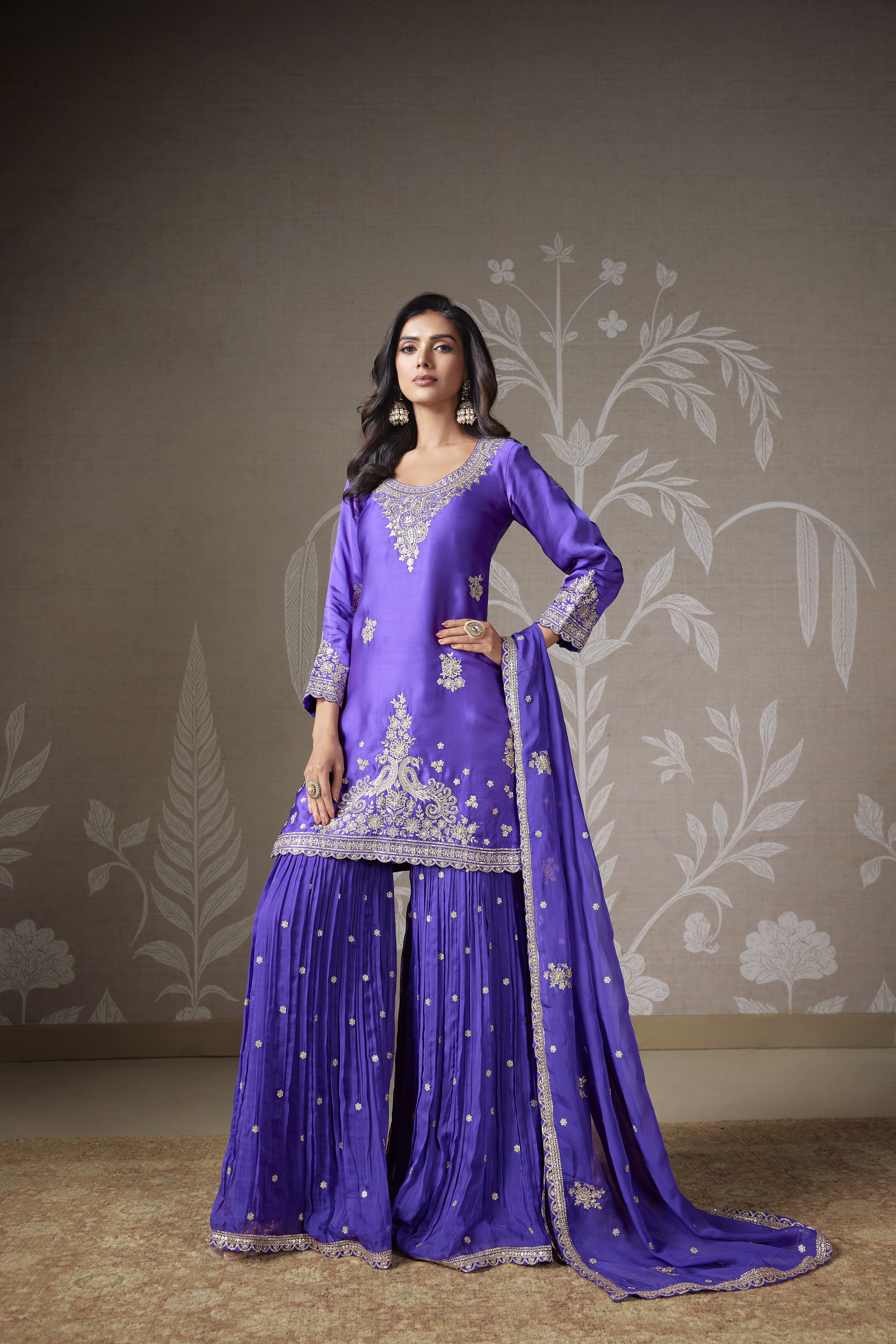 Woman wearing a purple embroidered sharara suit with matching dupatta and pleated sharara pants featuring intricate zari detailing.