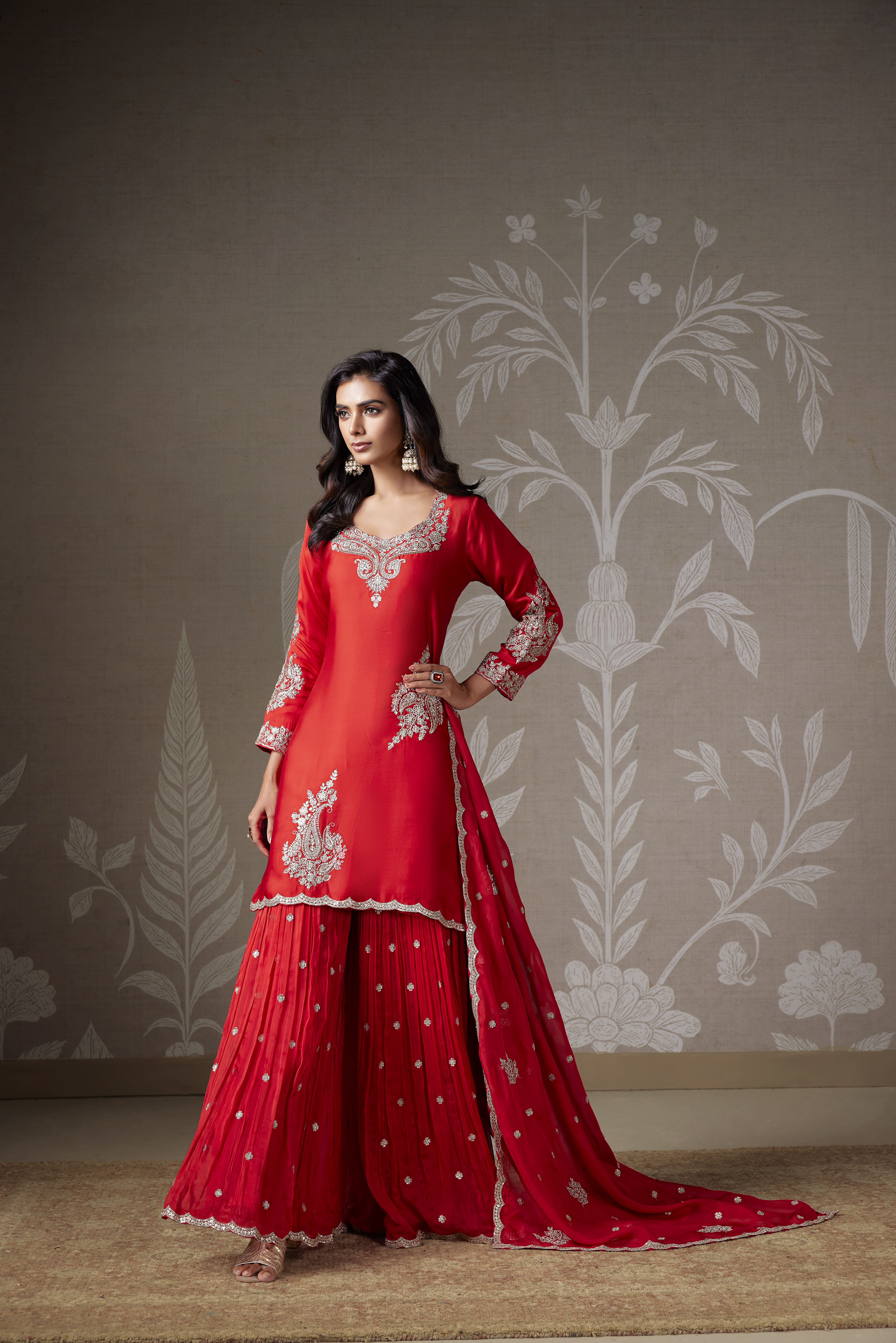 Model wearing a red embroidered sharara set with a matching dupatta and silver detailing