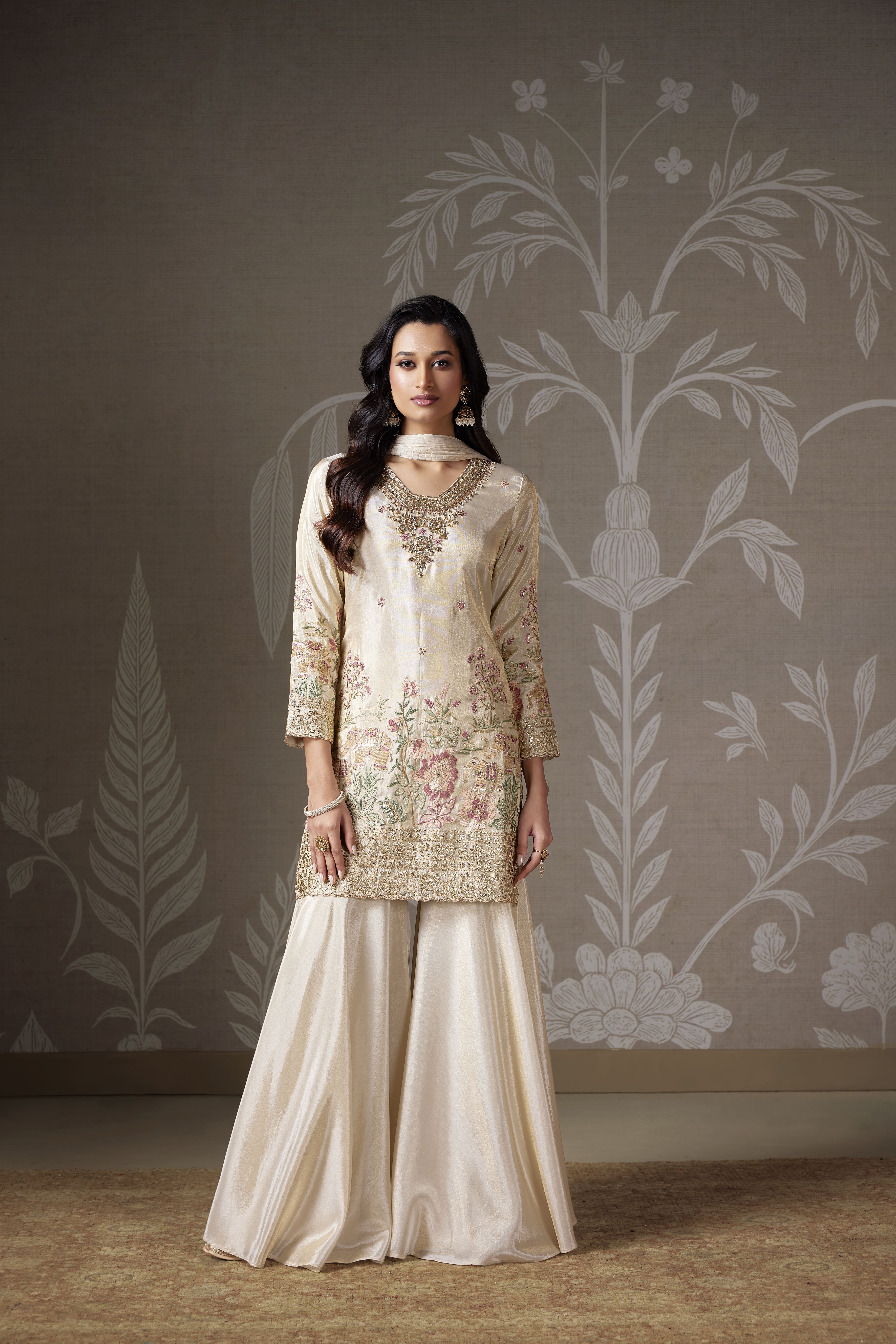 A woman posing in an elegant ivory-cream silk-satin sharara suit with intricate floral embroidery and gold zari details on the neckline and hem, set against a subtle damask-patterned background.