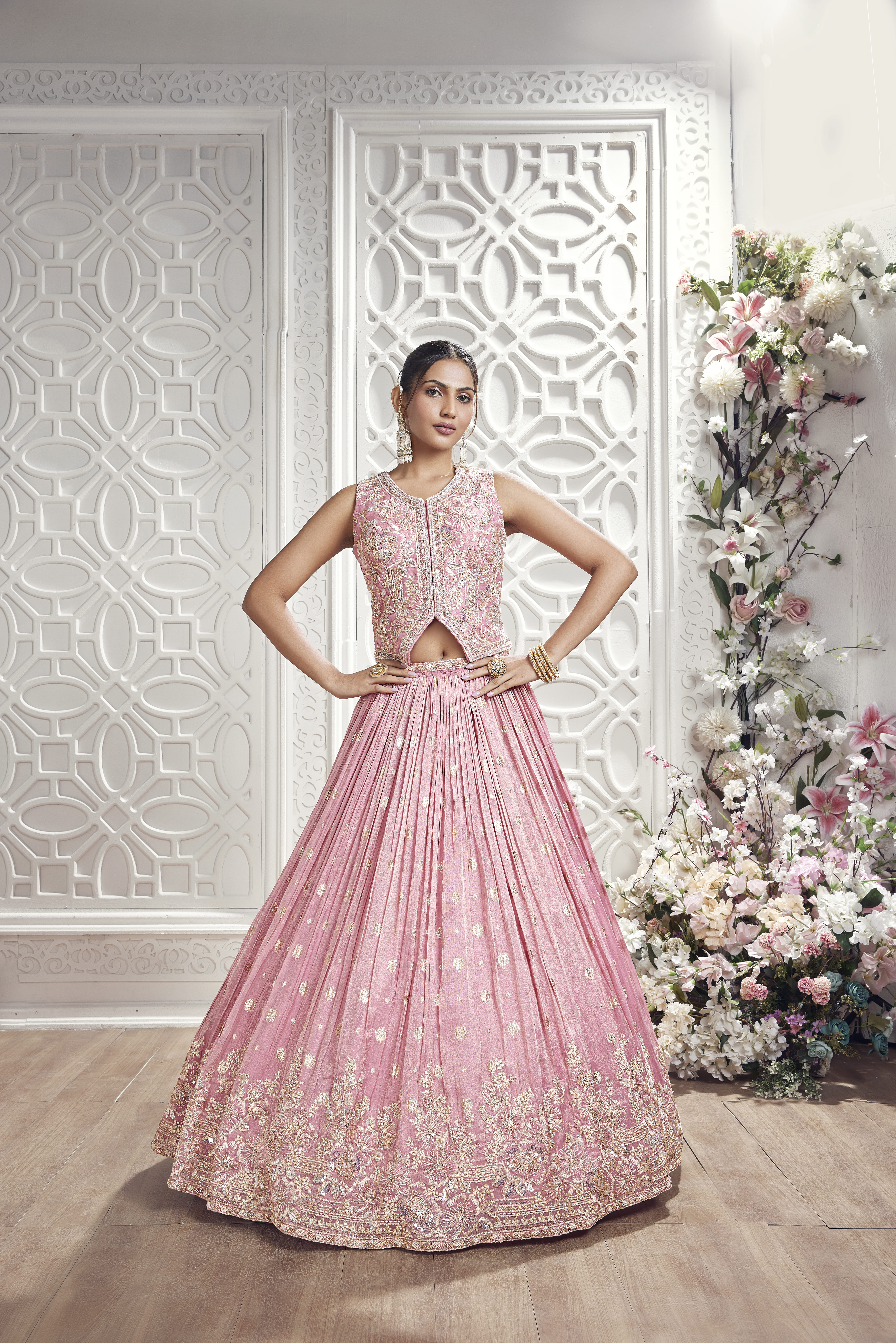 A woman modeling a blush pink ethnic set featuring a sleeveless embroidered peplum top with a front slit and a pleated lehenga skirt with a gold embroidered border, standing against a white carved wall backdrop.