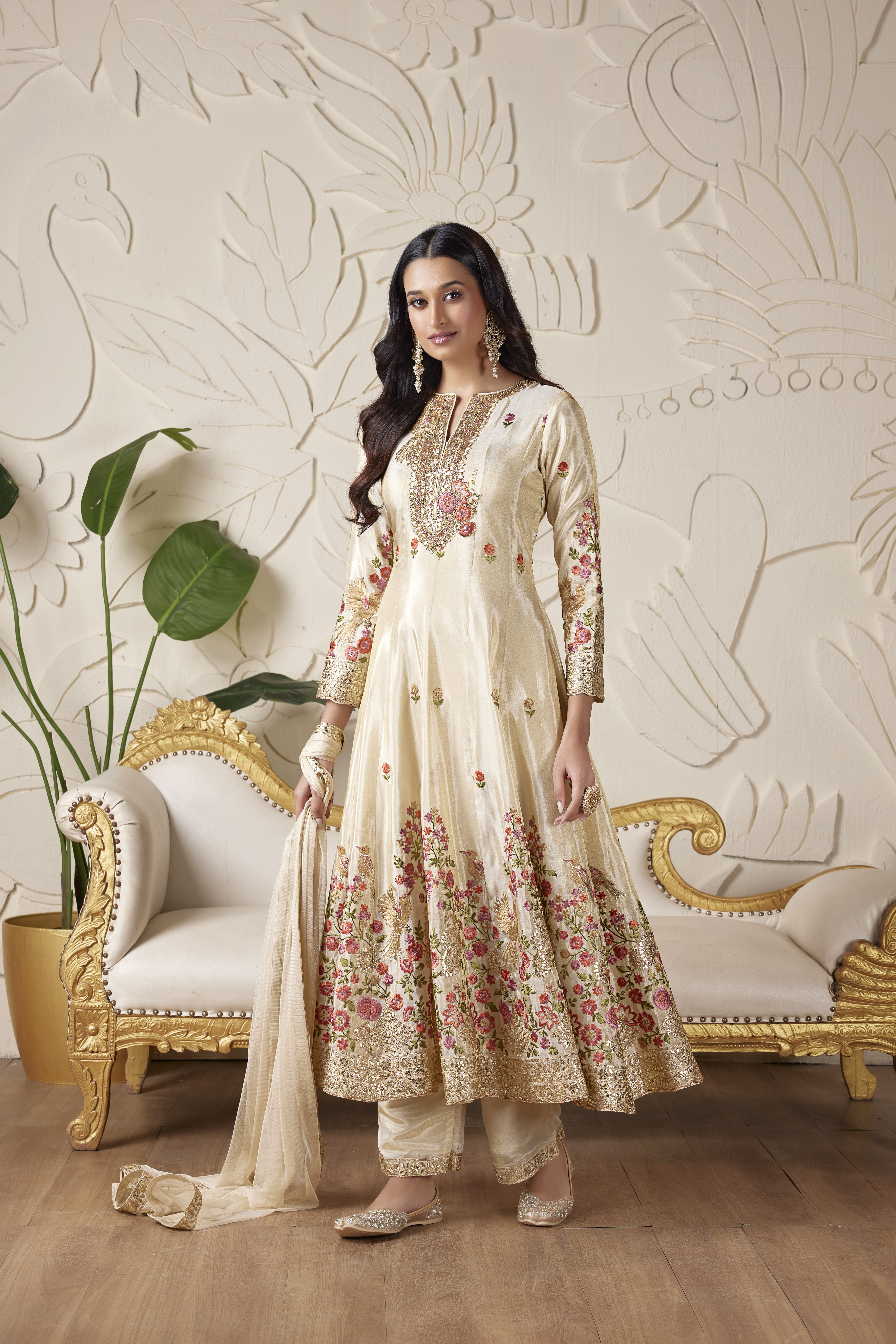 A woman modeling a cream-ivory Anarkali suit with heavy pink floral embroidery along the flared skirt hem and gold zari work on the neckline, holding a matching dupatta beside a vintage white and gold chaise lounge couch.