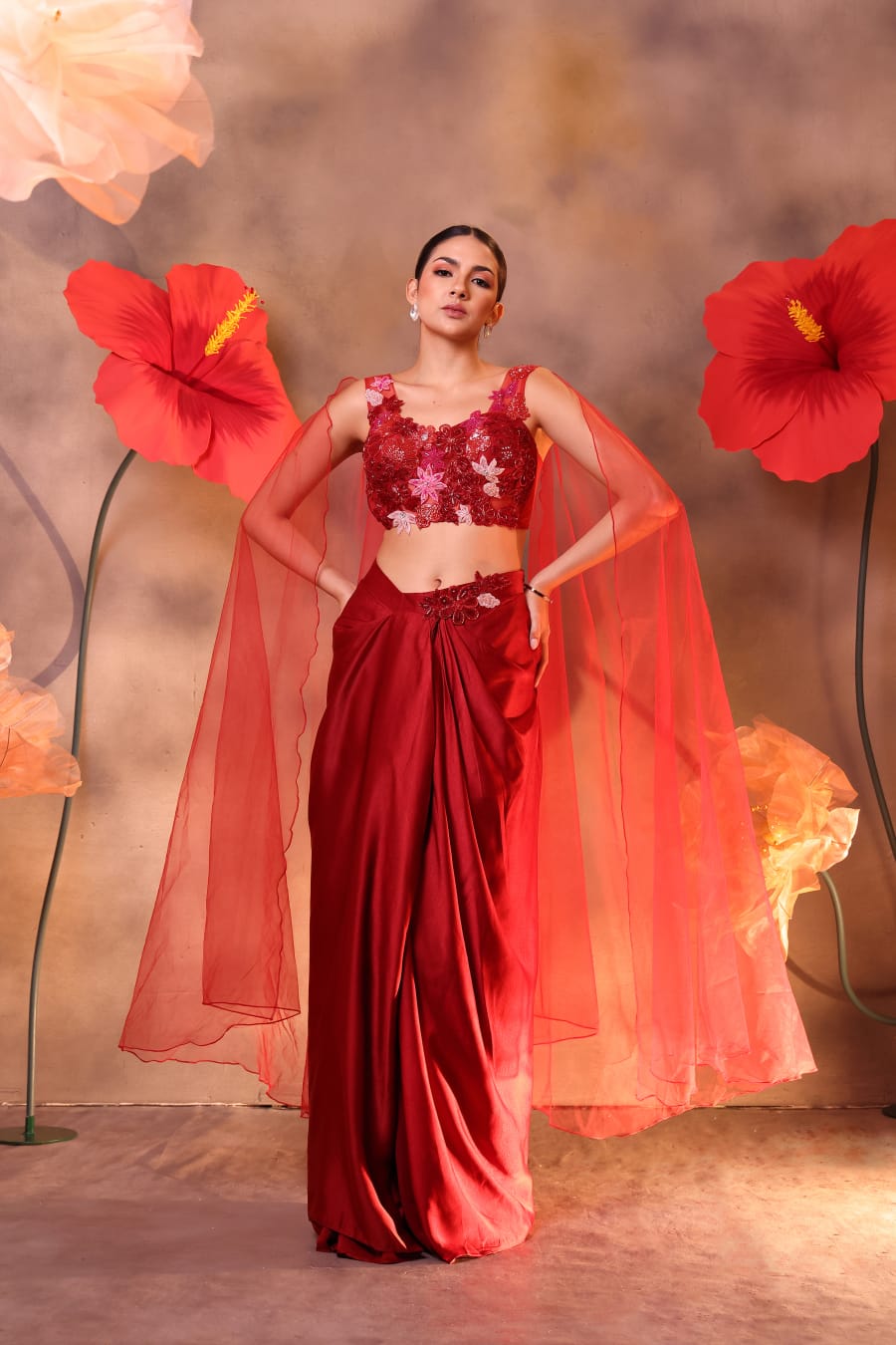 Woman wearing a ruby red embroidered blouse with attached sheer cape sleeves and a satin drape skirt, posing against a backdrop with large red flowers