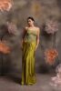 Woman wearing an olive green satin drape gown with a heavily embellished 3D floral corset top and spaghetti straps, posing against a backdrop with large pastel flowers