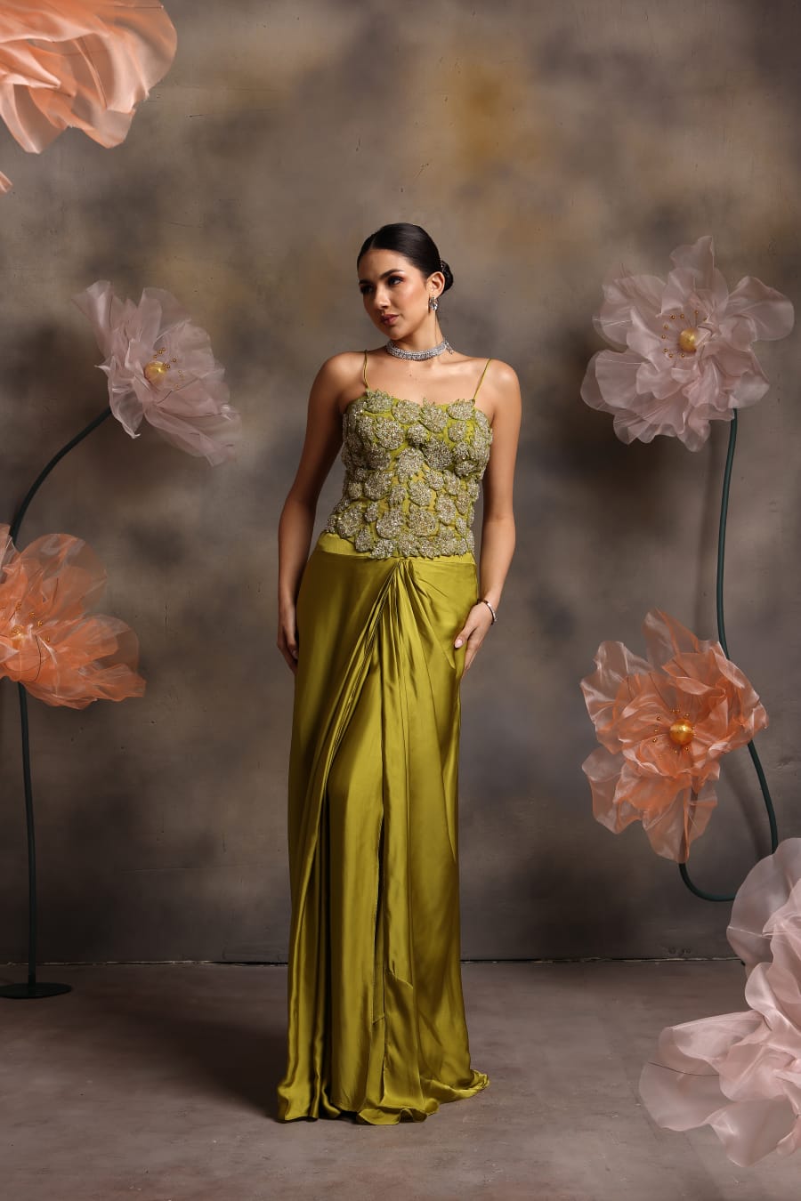 Woman wearing an olive green satin drape gown with a heavily embellished 3D floral corset top and spaghetti straps, posing against a backdrop with large pastel flowers