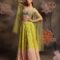 Woman wearing a lime yellow to peach ombre crushed lehenga with multicolor embroidered blouse and sheer net cape sleeves, posing against a backdrop with large pastel flowers