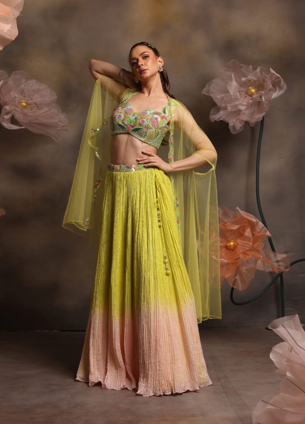Woman wearing a lime yellow to peach ombre crushed lehenga with multicolor embroidered blouse and sheer net cape sleeves, posing against a backdrop with large pastel flowers