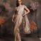 Woman wearing a champagne gold pre-draped satin saree with crystal border and a silver embellished halter-neck blouse, posing against a floral backdrop