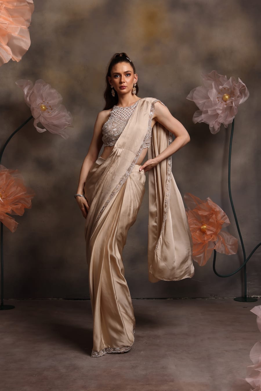 Woman wearing a champagne gold pre-draped satin saree with crystal border and a silver embellished halter-neck blouse, posing against a floral backdrop