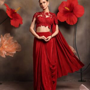 Woman wearing a red lace blouse with cut-out detailing and attached cape, paired with a pre-draped satin saree gown, standing against a floral backdrop