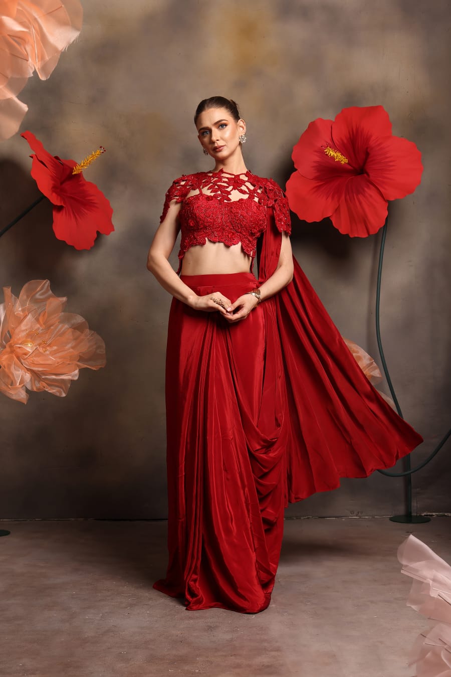 Woman wearing a red lace blouse with cut-out detailing and attached cape, paired with a pre-draped satin saree gown, standing against a floral backdrop