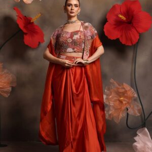 Woman wearing a burnt orange satin drape skirt with multicolor embellished bustier and embroidered cape, posing against a floral backdrop
