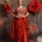 Woman wearing a burnt orange satin drape skirt with multicolor embellished bustier and embroidered cape, posing against a floral backdrop
