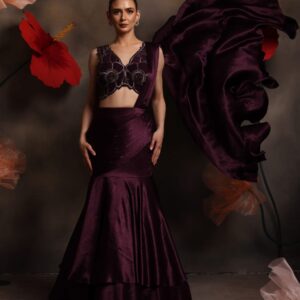 Woman wearing a plum satin fishcut drape saree gown with silver embellished sleeveless blouse and flowing pallu, posing against a floral backdrop