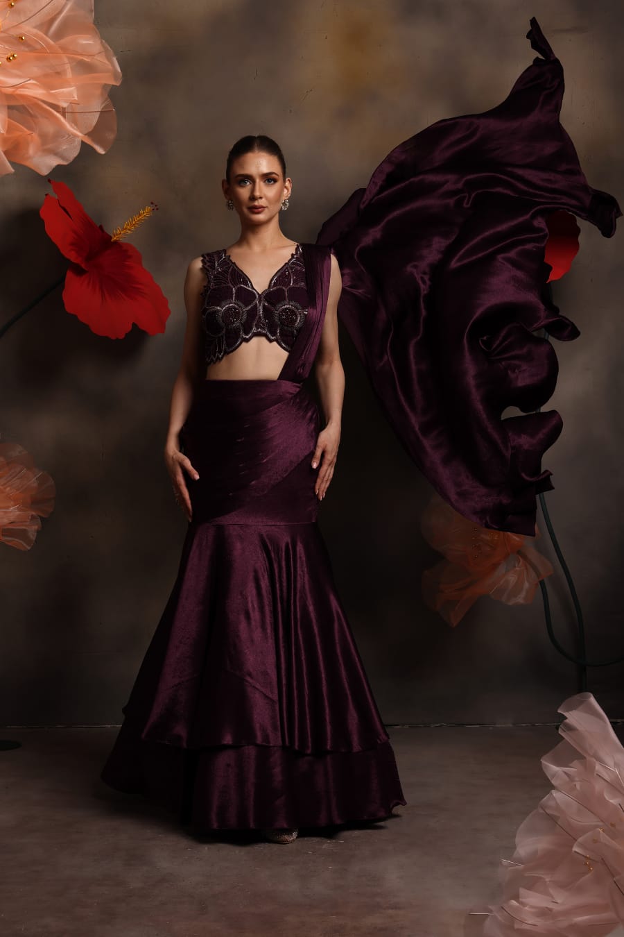 Woman wearing a plum satin fishcut drape saree gown with silver embellished sleeveless blouse and flowing pallu, posing against a floral backdrop