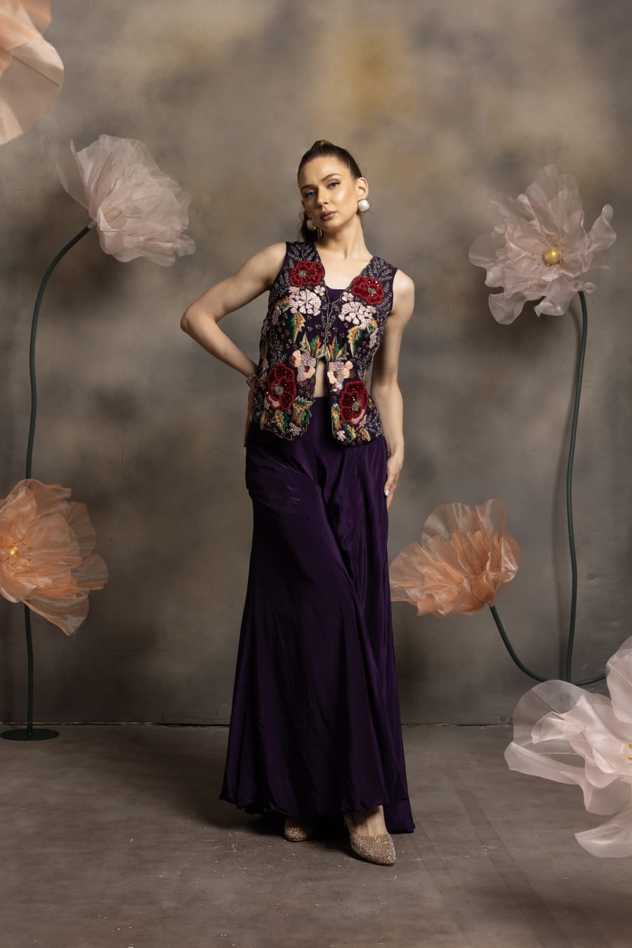 Woman wearing an aubergine purple flared pants set with matching bustier and a sleeveless jacket featuring 3D floral embroidery in red and pink, posing against a floral backdrop