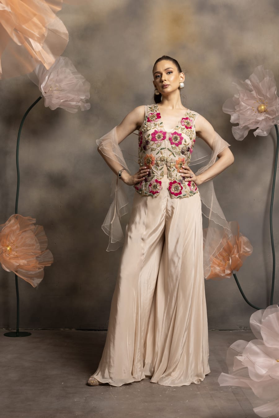 Woman wearing an ivory satin palazzo set with a sleeveless jacket featuring 3D floral embroidery in pink and orange, and a sheer organza dupatta, posing against a floral backdrop