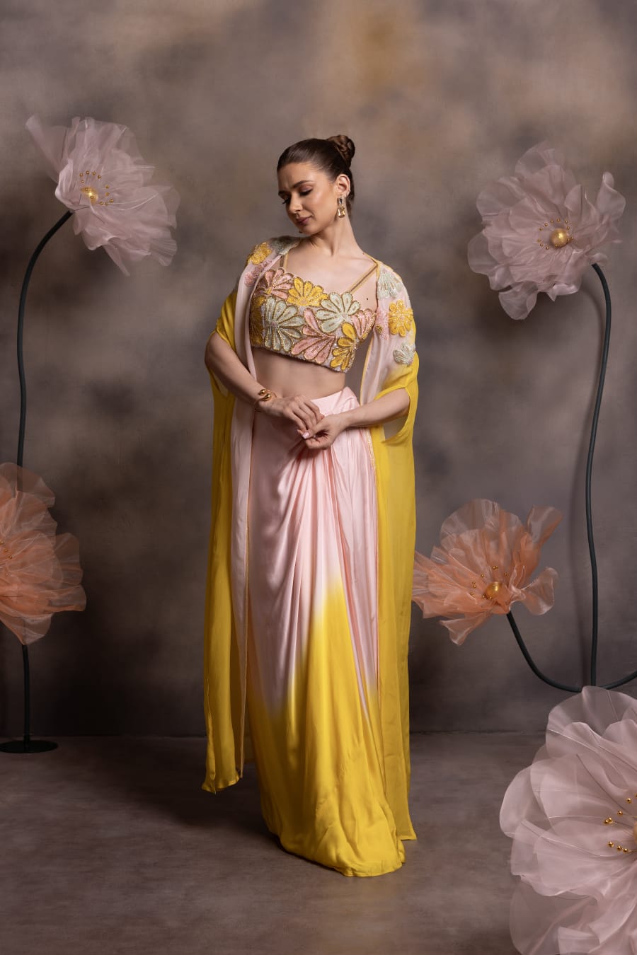 Woman wearing a blush pink to yellow ombre satin drape skirt with 3D floral embroidered bustier and matching cape, posing against a floral backdrop