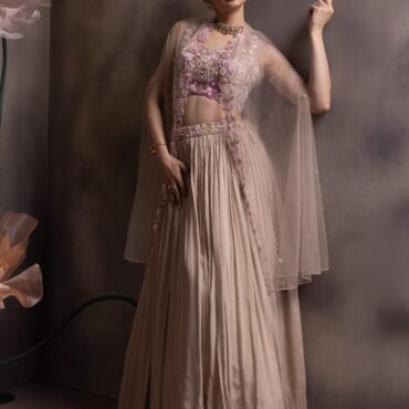 Woman wearing a taupe pleated lehenga with a lilac embroidered crop top and sheer net jacket with floral borders, posing against a muted floral backdrop