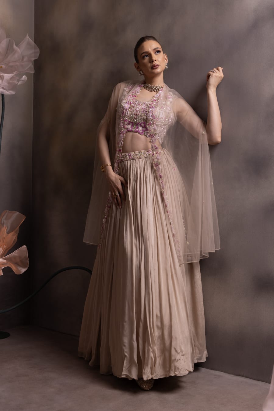 Woman wearing a taupe pleated lehenga with a lilac embroidered crop top and sheer net jacket with floral borders, posing against a muted floral backdrop
