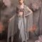 Woman wearing a silver-grey satin drape skirt with lavender mirror-work bustier and a sheer organza cape with crystal tassel embroidery, posing against a floral backdrop