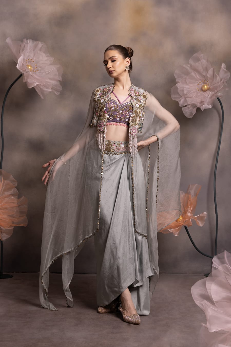 Woman wearing a silver-grey satin drape skirt with lavender mirror-work bustier and a sheer organza cape with crystal tassel embroidery, posing against a floral backdrop