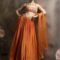 Woman wearing a tangerine orange pleated silk lehenga with multicolor embroidered halter blouse and sheer net dupatta, posing against a muted backdrop