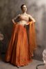 Woman wearing a tangerine orange pleated silk lehenga with multicolor embroidered halter blouse and sheer net dupatta, posing against a muted backdrop