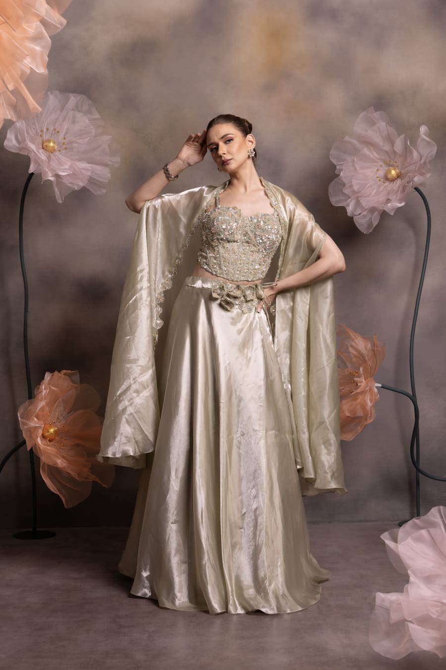 Woman wearing a metallic champagne tissue lehenga with crystal-embellished corset blouse and scalloped cape, posing against a floral backdrop
