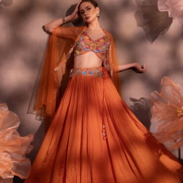 Woman wearing a rust-orange pleated lehenga with multicolor embroidered blouse and sheer organza cape, posing against a floral backdrop