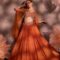 Woman wearing a rust-orange pleated lehenga with multicolor embroidered blouse and sheer organza cape, posing against a floral backdrop