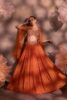 Woman wearing a rust-orange pleated lehenga with multicolor embroidered blouse and sheer organza cape, posing against a floral backdrop