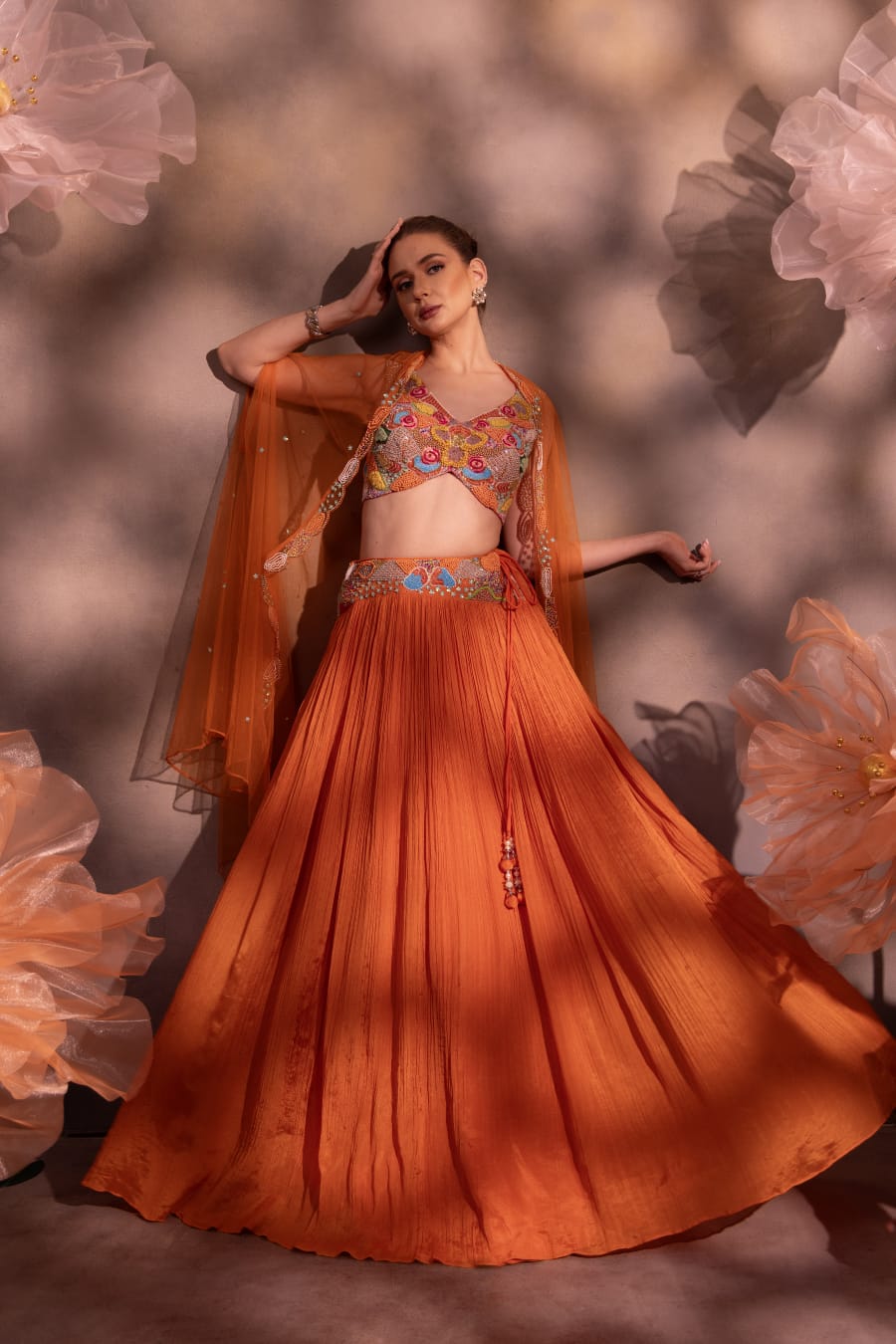 Woman wearing a rust-orange pleated lehenga with multicolor embroidered blouse and sheer organza cape, posing against a floral backdrop